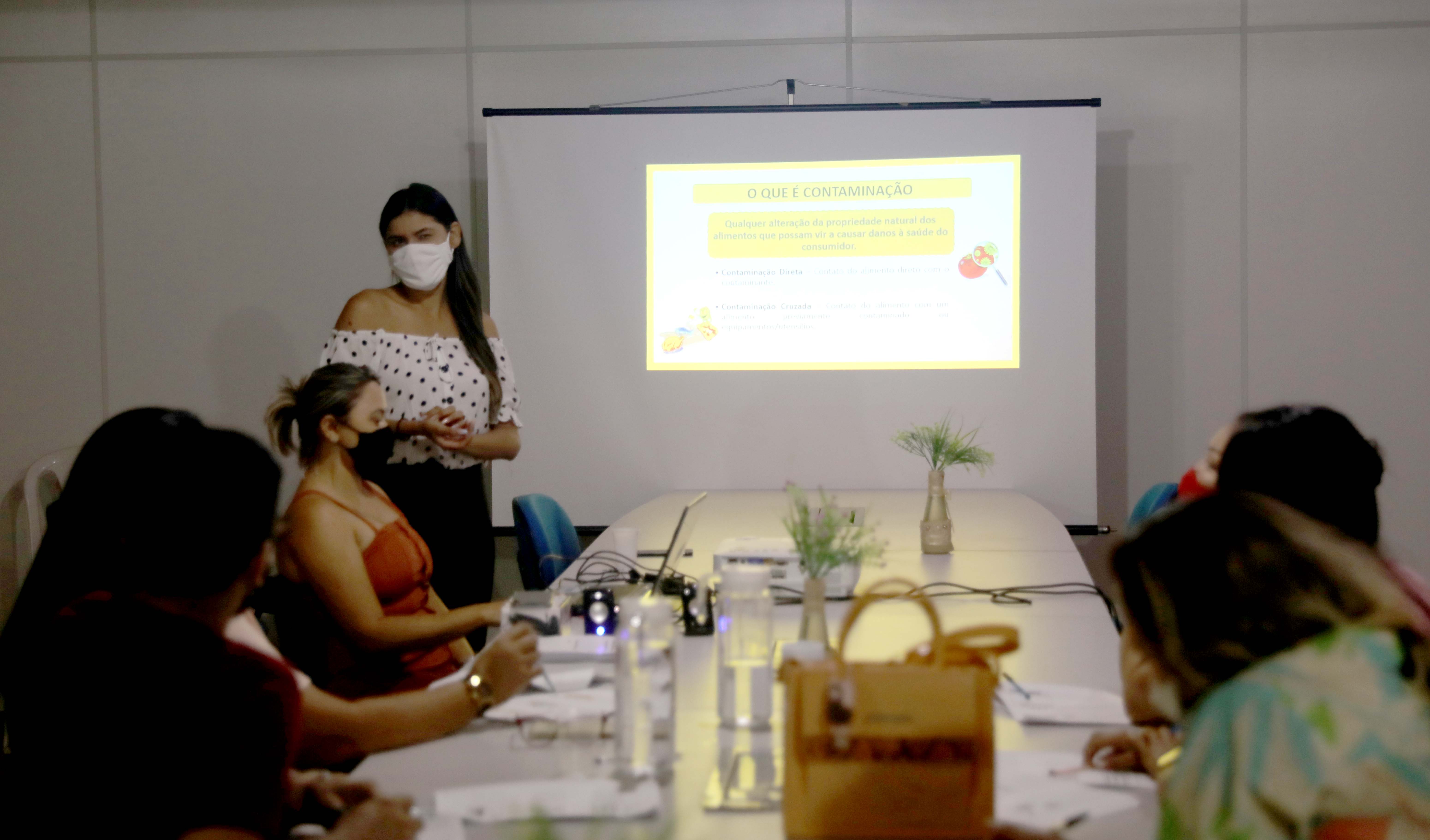 a foto mostra uma instrutora na frente de um telão enquanto dá aula. Os alunos estão sentados em uma mesa comprida